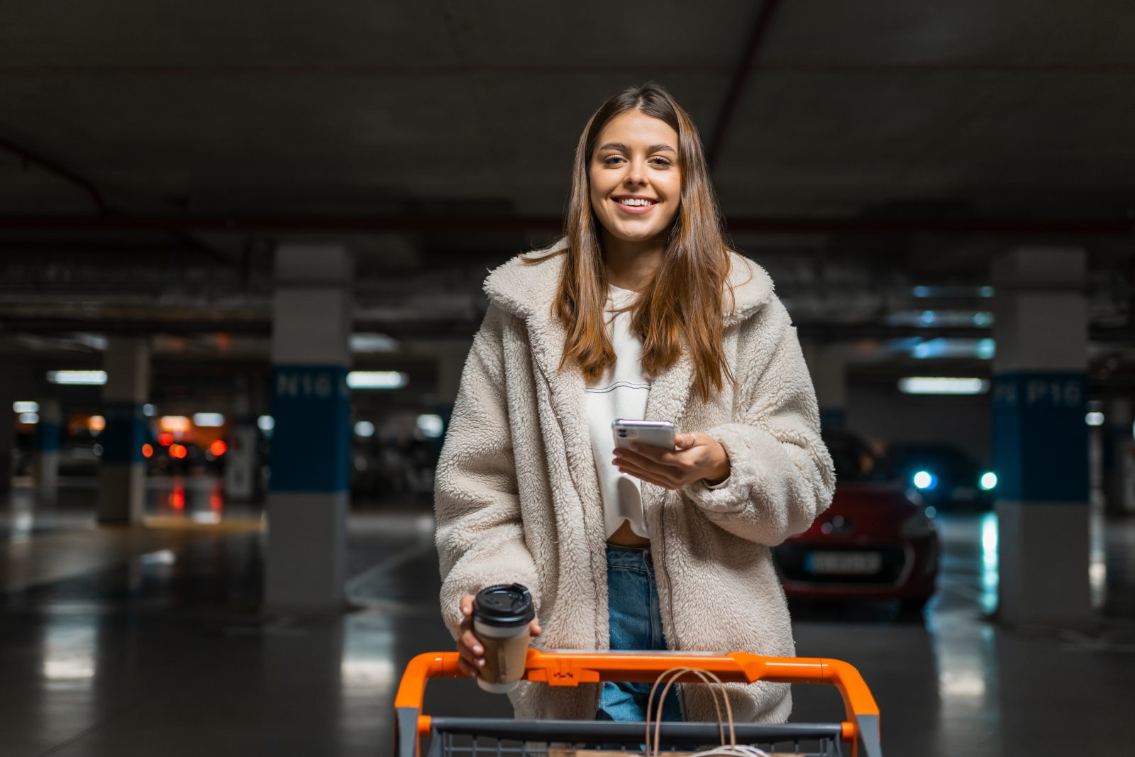 Pretty young woman with smartphone in hands and shopping trolley in the underground parking of shopping mall. Portrait of female shopper with shopping cart